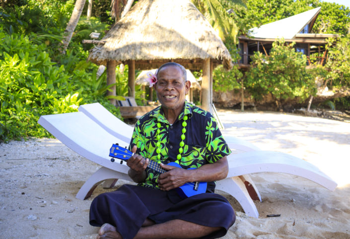 fijian man sitting on a beach smiling and holding ukelele